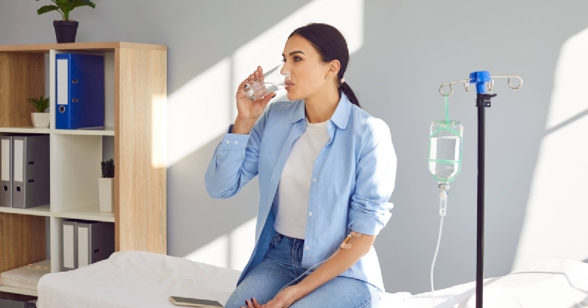 A woman sits on a medical bed receiving IV infusion therapy while drinking a glass of water in Katy TX